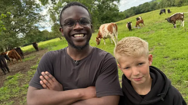 A man with black hair, black glasses and a black t-shirt has his arms folded and is smiling at the camera. Next to him is a young boy with blonde hair wearing a black hoodie. They are standing in a field with grass and cows behind them