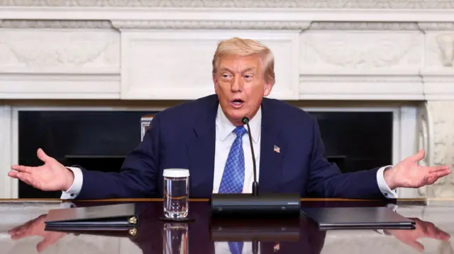 Donald Trump sitting down at a wooden desk with his arms open to the sides. He's speaking into a thin mic whilst sitting in front of a marble fireplace