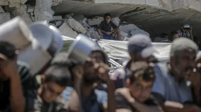 A boy (rear) sitting among the rubble of a damaged building looks on as internally displaced Palestinians gather to receive food from a charity kitchen