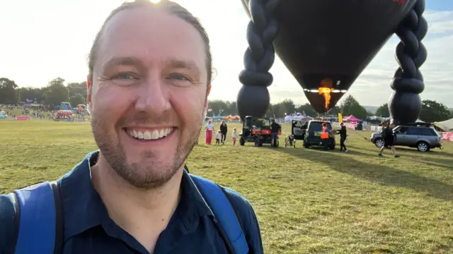 Ross Pollard standing on a grassy field a Ashton Court with a large black hot air balloon being prepared for take-off