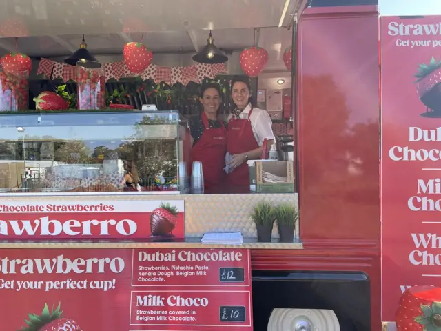 Jay Graham is on the left standing next to Caroline Morley. They are both wearing red aprons and are standing isnide of their strawberry business food truck