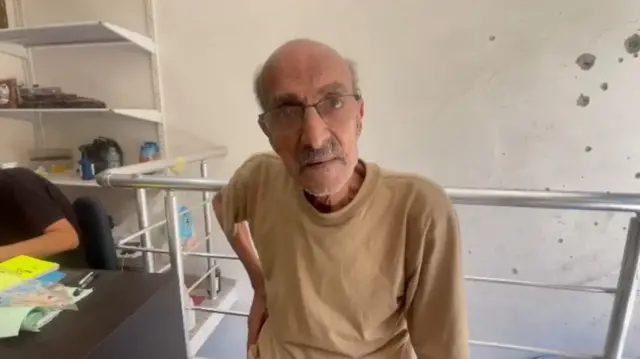 Elderly man with glasses and grey hair to the side of his otherwise bald head looks into the camera. He's standing insider a white-walled room with what appears to be bullet or shrapnel damage to the right. He's in a beige shirt, his right hand on his hip, there's another person in a short-sleeved black shirt sitting at a desk to the man's right