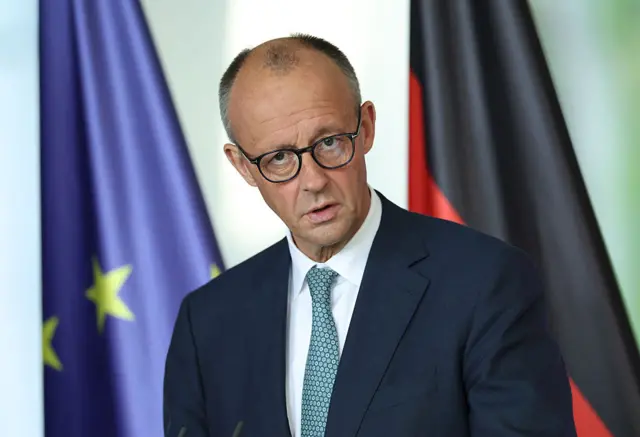 German Chancellor Friedrich Merz speaking at a news conference. He is wearing a dark suit and glass. In the background the EU and German flags are visible.