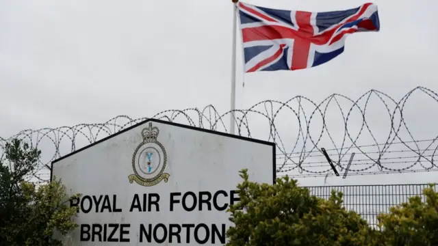 The sign in front of RAF Brize Norton with a barbed wire fence and a union flag behind it