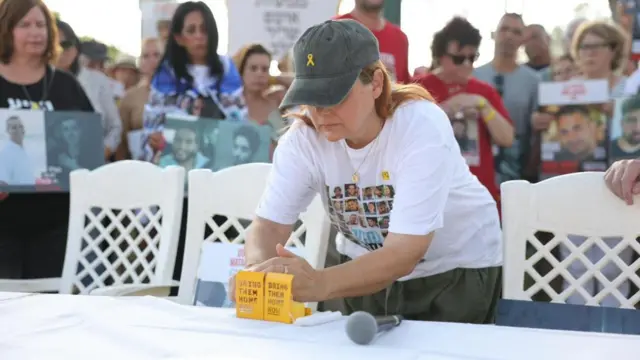 A woman leans forward over a table with empty chairs, lighting candles in boxes saying 'bring them home', surrounded by families of hostages being held by Hamas