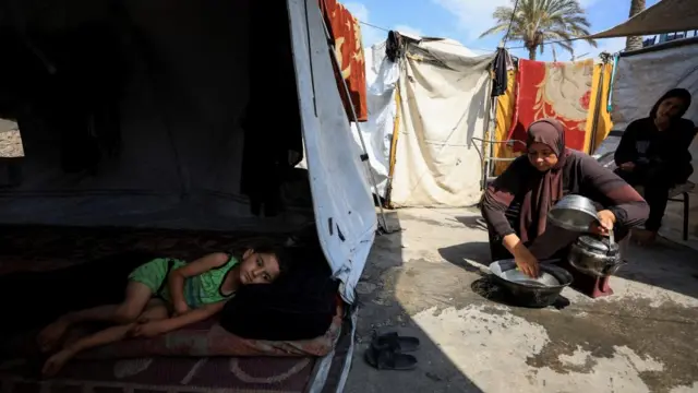 A young child lies in a tent in the shade in Gaza City while a woman crouches in the sun on the concrete, cooking in a pot with another woman sitting on a chair watching her
