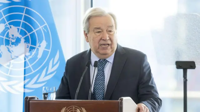 Guterres speaks into a microphone that is attached to a stand on a podium. He holds the podium on its side with one hand. Behind him is a window and a UN flag.