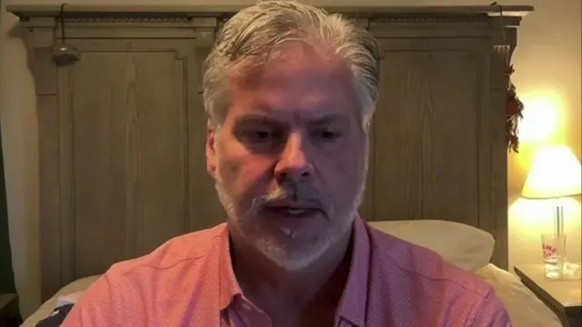 A man with grey hair and a pink shirt on sits in front of a bed