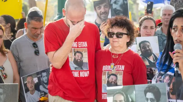 A man covers his face as be bowsn his head with emotion, standing in a group of hostage families holding placards with pictures of the missing