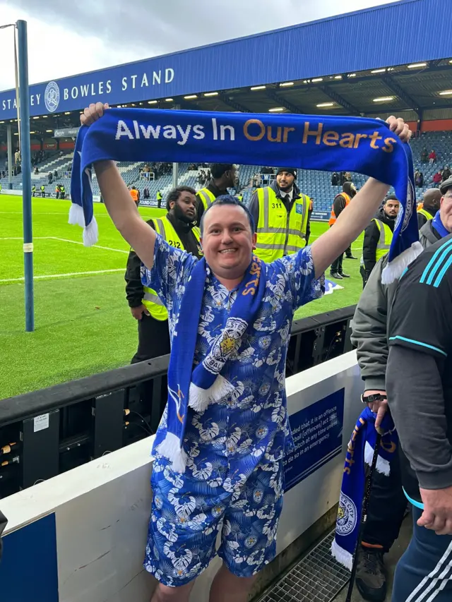 Man dressed in Leicester City gear holding up scarf inside ground