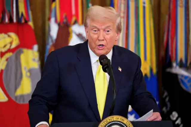President Donald Trump speaks from a lectern during a news conference. He is speaking behind a lectern bearing the presidential seal and wearing a yellow tie and US flag pin.