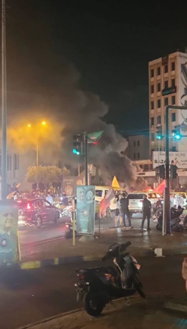 A still from a video showing tyre fires in Beirut. In the foreground is a parked motorbike. People are gathered on a nearby traffic island as smoke billows in the air in the distance from a fire