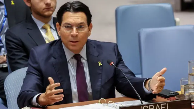 Danny Danon sits at wooden desk during UN Council meeting. He's wearing a dark suit, a yellow ribbon pin on his left lapel, another man partially visible sitting on a blue chair behind him