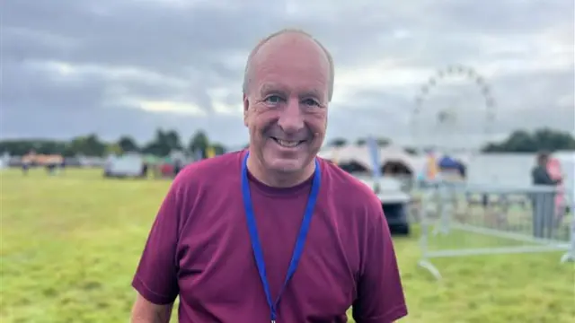 Clive Bailey wearing a burgundy t-shirt with a blue lanyard around his neck standing in Ashton Court Estate