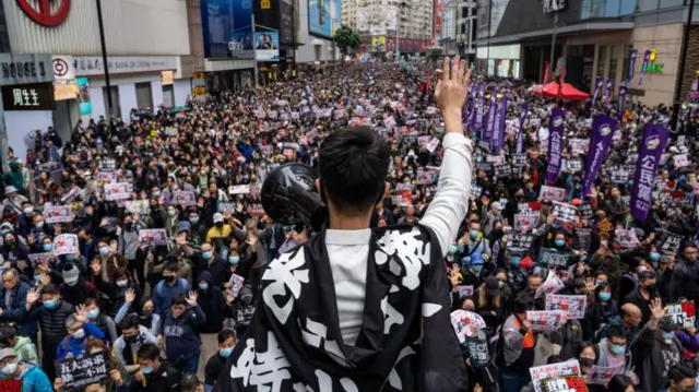 Backview of a man draped in a black banner with revolutionary slogan, holding a loudspeaker. In front of him is a large crowd on a street
