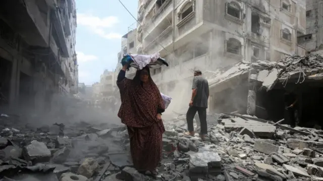 A woman holds a tray on her head as she walks through a destroyed street, full of rubble, in Gaza City.