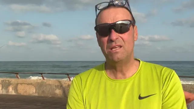 A man wearing sunglasses and running gear speaks in front of a Tel Aviv beach