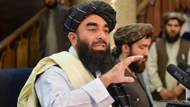 A man in a white shirt, cream sash and black turban speaks into a microphone