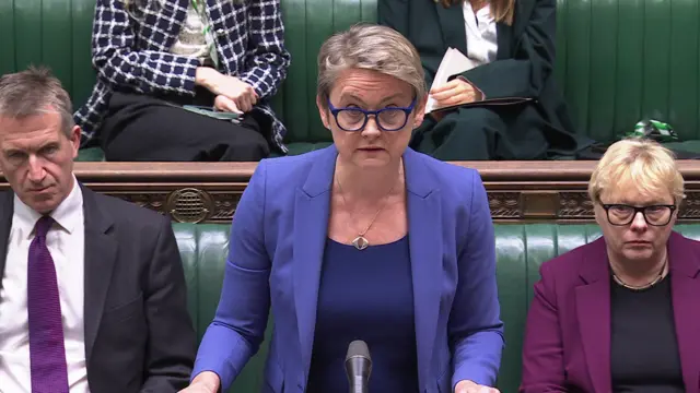 Cooper stands speaking at the dispatch box in the House of Commons, wearing a blue blazer, blue glasses and blue shirt.