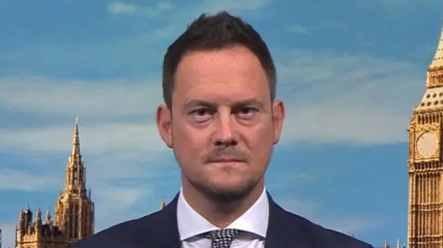 Headshot of Morgan who sits looking stern with his mouth closed looking into the camera. He wears a suit and tie, and sits in front of a background which shows blue sky, big ben, and the top of the Parliament buildings