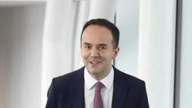 Headshot of Murray who smiles, wearing a suit and red tie, in a room with white walls