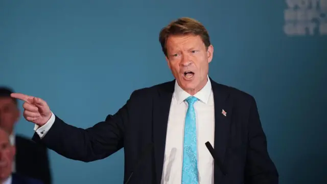 Tice speaks into two small microphones on stands, with his mouth open while he raises one hand and points to the side