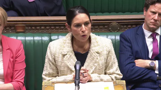 Priti Patel in a cream jacket at the dispatch box in front of the green benches of the House of Commons
