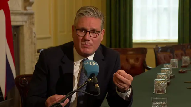 Starmer gestures with both hands as he speaks int a blue 5Luve microphone. He sits in the cabinet room in Downing St
