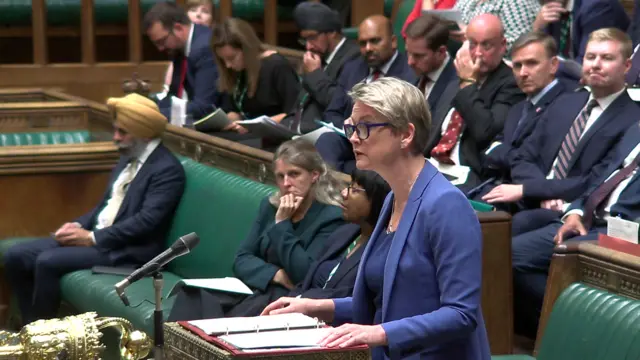 A side view of Cooper at the dispatch box. Behind her, MPs sit on the benches. Some watch her, others look at notes