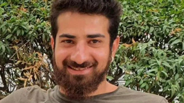 Idan Shtivi smiling and wearing a grey top in front of a leafy hedge