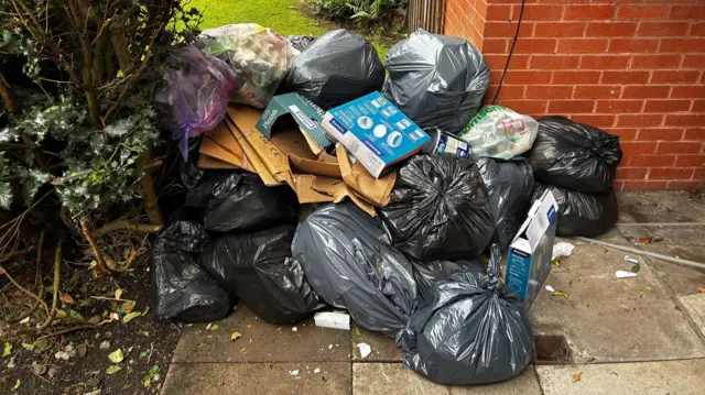 A pile of black bin bags and cardboard boxes in a pile on the ground next to a red brick building and a bush