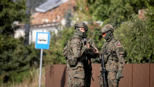 Three armed forces members standing a huddle, holding large guns. One looks at the camera