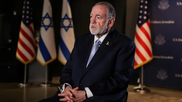 Mike Huckabee sits on a chair wearing a suit with US and Israeli flags in the background.