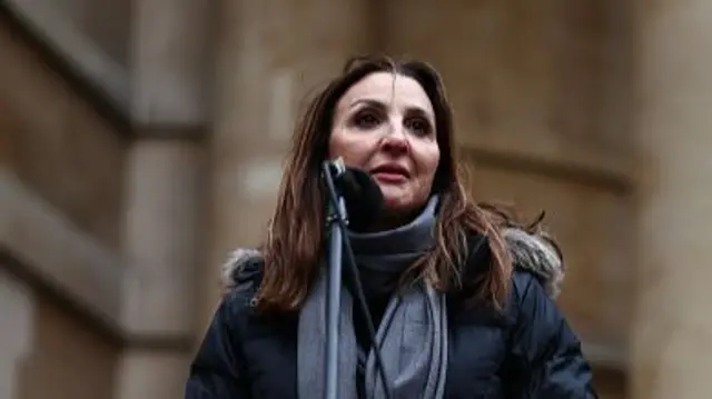 Fleur Hassan-Nahoum stands by a microphone wearing a coat and scarf
