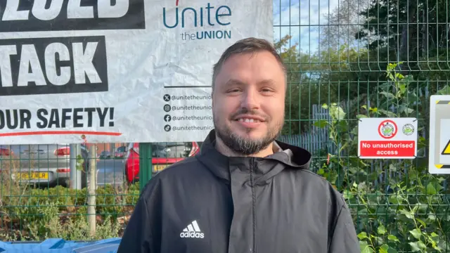 A man with short brown hair is wearing a black adidas waterproof jacket. He is standing in front of a tall green metal fence. Attached to the fence is a large banner that has the "unite the union" logo on it