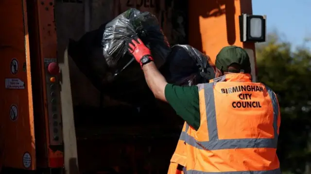 A man is wearing an orange hi-vis jacket that says "birmingham city council" on the back. He is wearing a green cap and red gloves, and is putting two black bin bags into the back of an orange waste lorry