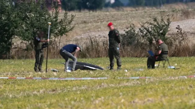 A group of officials standing in a field looking at a drone on the ground