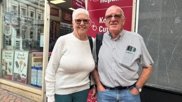 A man and a woman are standing in the street next to each other outside a shop. The man has very short white hair with tinted glasses, and is wearing a white and grey checked shirt. The woman has short white hair and is wearing tinted glasses, and is wearing a white jumper