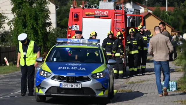 A police car parked on a street in front of a fire engine. There's a lot of firemen and some other emergency workers walking and standing around