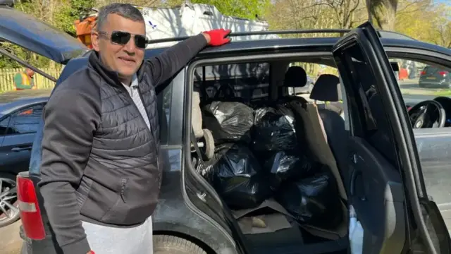A man with short black and grey hair is wearing sunglasses, a black puffer jacker and grey trousers, as well as red gloves. He has piled black bin bags inside his car, which he is holding onto with the door open