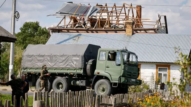 A military truck parked in front of a house with a destroyed roof, with all the beams exposed. It is day outside