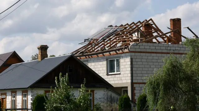 A house in the foreground is unscathed. the one in the back has a damaged roof - all the beams are showing