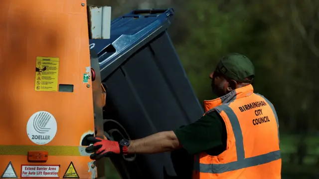 A man with a green hat is wearing an orange hi-vis that says "birmingham city council" on the back. He is wearing red and black gloves and is holding a black wheeely bin to the back of an orange waste lorry