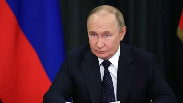 Putin wearing a suit. He sits in front of a Russian flag