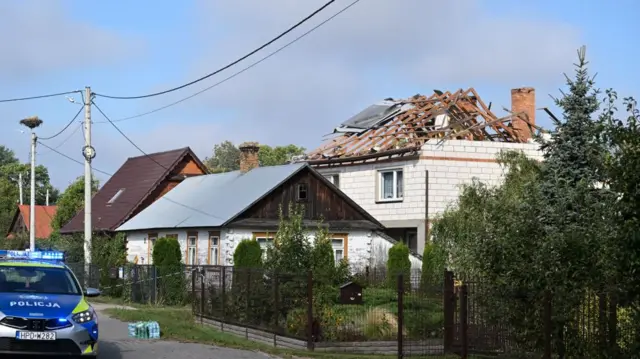 From the street, there is a house with a roof completely damaged and ripped apart. A smaller house sits in front of the damaged one, and they are  behind a metal fence. On the street a police car is parked. There's a lot of trees and thick green grass