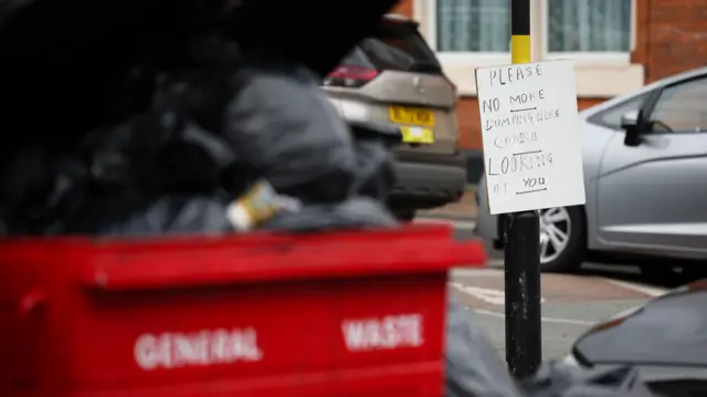 In the foreground is a red general waste bin filled and overflowing with black bin bags. In focus in the background is a sign attached to a lamppost that reads "Please no more dumping here. Camera looking at you"