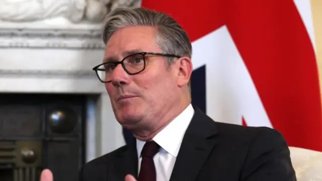 Starmer is sitting on a chair in front of a British flag and a fancy fireplace. He talks and gestures with both arms held up in front of him