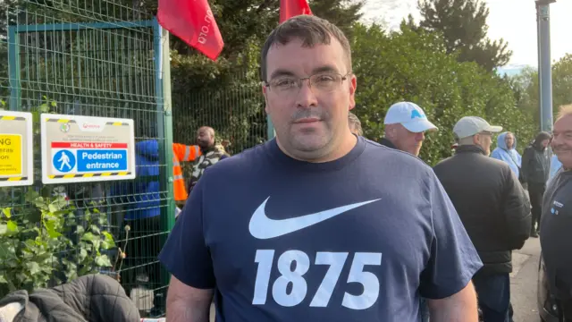 A man is standing on a picket line with a tall green fence in the background. He has brown hair and is wearing glasses. He is wearing a blue top with a large tick on it, and the it says "1875"