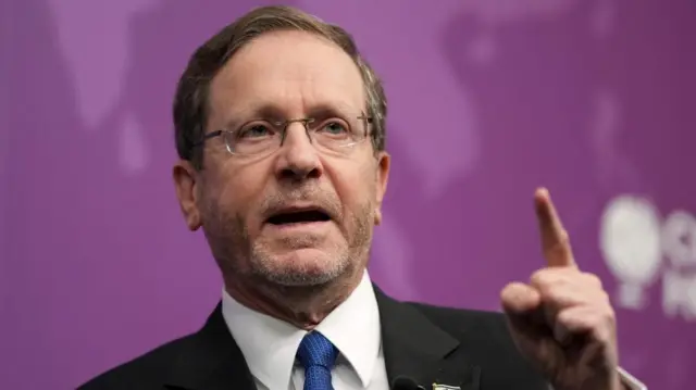 Headshot of Israeli President Isaac Herzog pointing his finger as he speaks. He wears glasses, a dark suit, white shirt and blue tie.