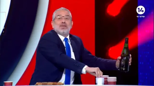 A still from the television programme shows Magal with an open bottle of champagne in one hand and reaching to grab a red plastic cup with the other
